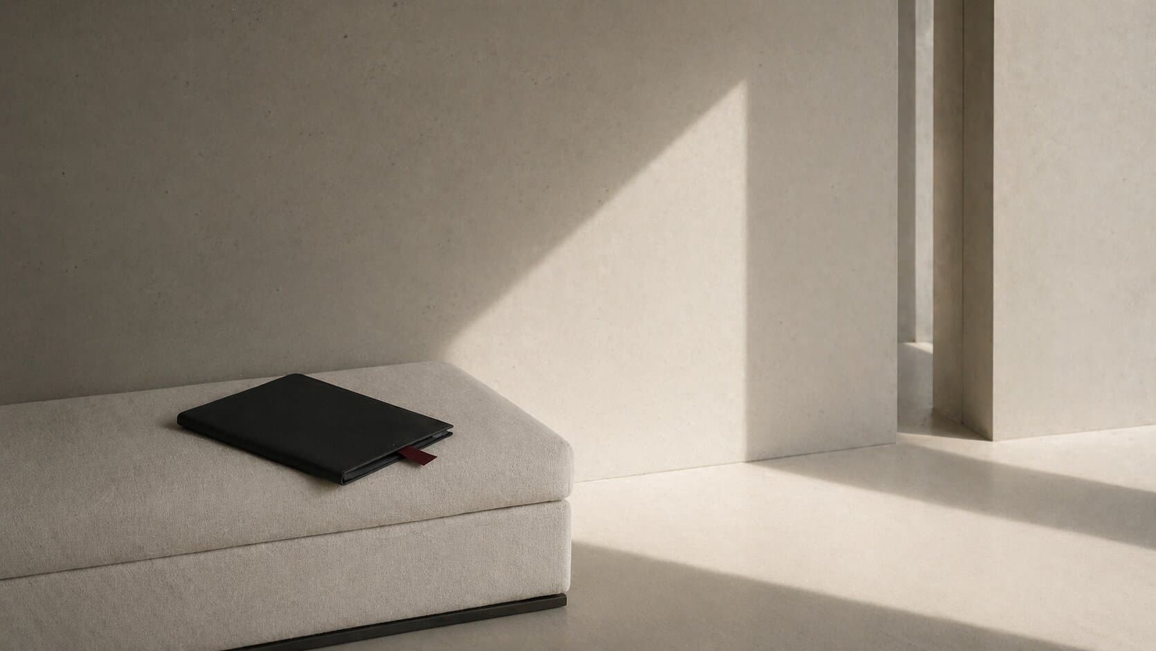 A matte-black folio on a bone-linen bench, late afternoon light cutting a long diagonal across a concrete wall.