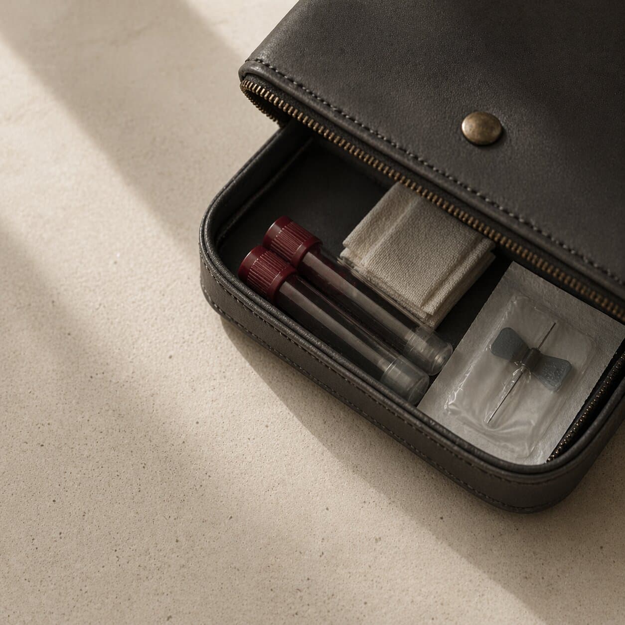 A soft black leather collection kit opened on a bone-cream stone surface — oxblood-capped vials, a gauze pad, and a sealed butterfly needle visible inside. Hard afternoon light cuts a clean diagonal shadow.